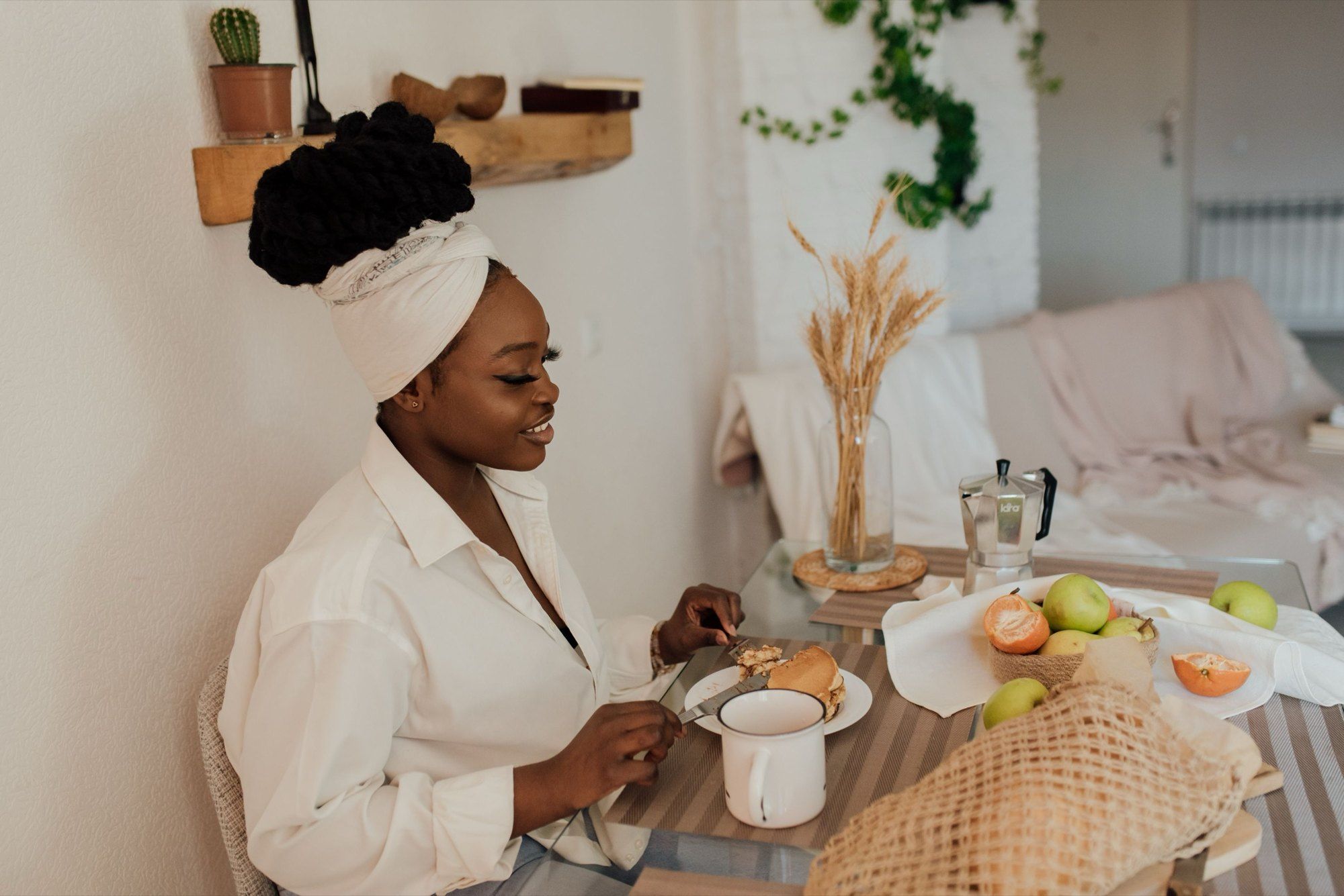 A woman in a head wrap having breakfast with fruit and tea at home, in soft window light.