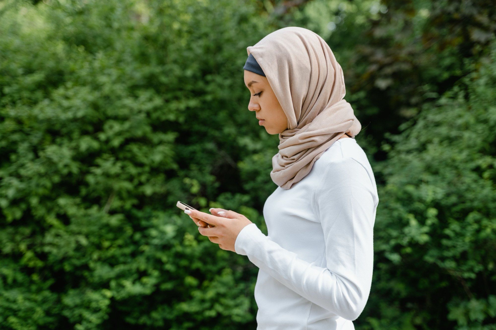 A woman in hijab looking at her phone outdoors against greenery.